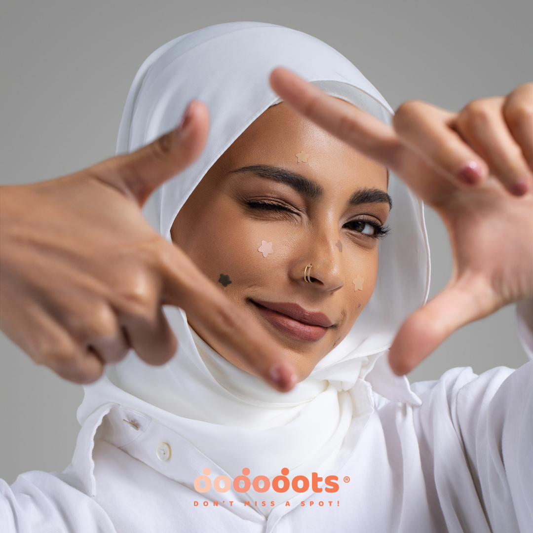 Woman in a white hijab winking through a finger frame, wearing skin-tone star-shaped Dododots pimple patches on her cheek and forehead